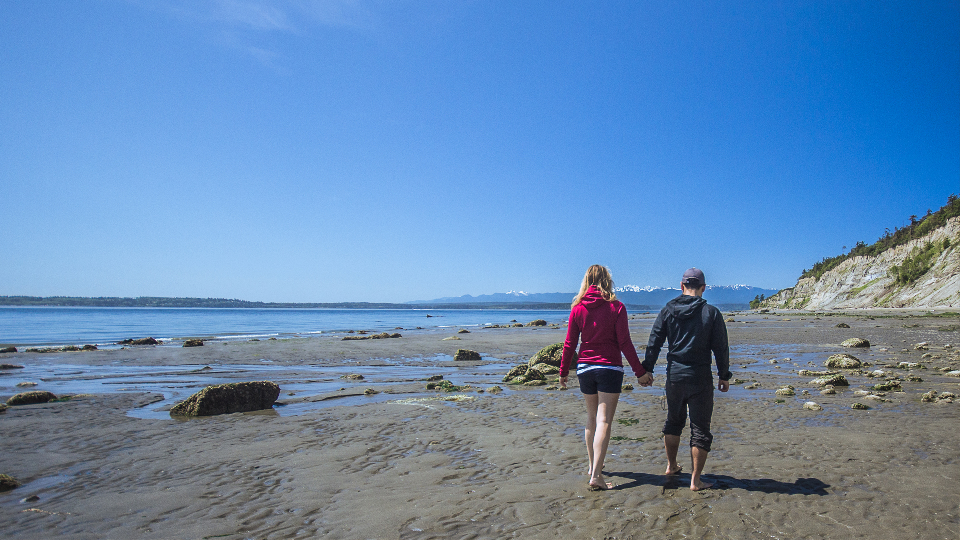 Ein Paar genießt einen Spaziergang am Strand von Tossens. Im Hintergrund erstreckt sich die Nordsee, während die frische Meeresbrise für eine entspannte und harmonische Urlaubsatmosphäre sorgt.